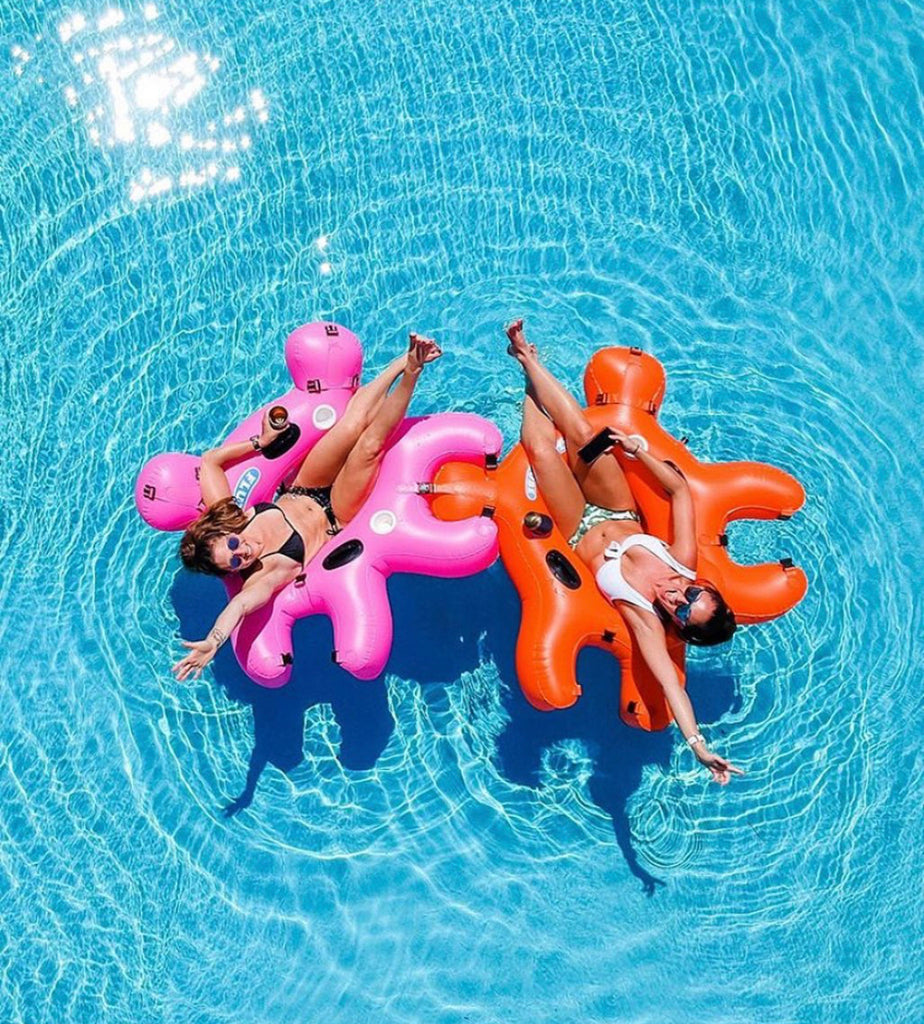 Two ladies in bathing suites floating in the pool on Orange and Pink Interlocking Fluzzle Tube floaties. 