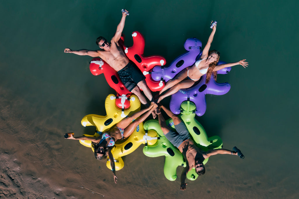 4 people in bathing suite floating in connecting Red, Green, Purple and Yellow Fluzzle Tubes puzzle shaped inflatable and interlocking river, lake or pool tube with inflatable back rest, mesh bottom, expandable cup holders, 2 durable handles. 12 nylon connectors. Interlocking floating cooler. 