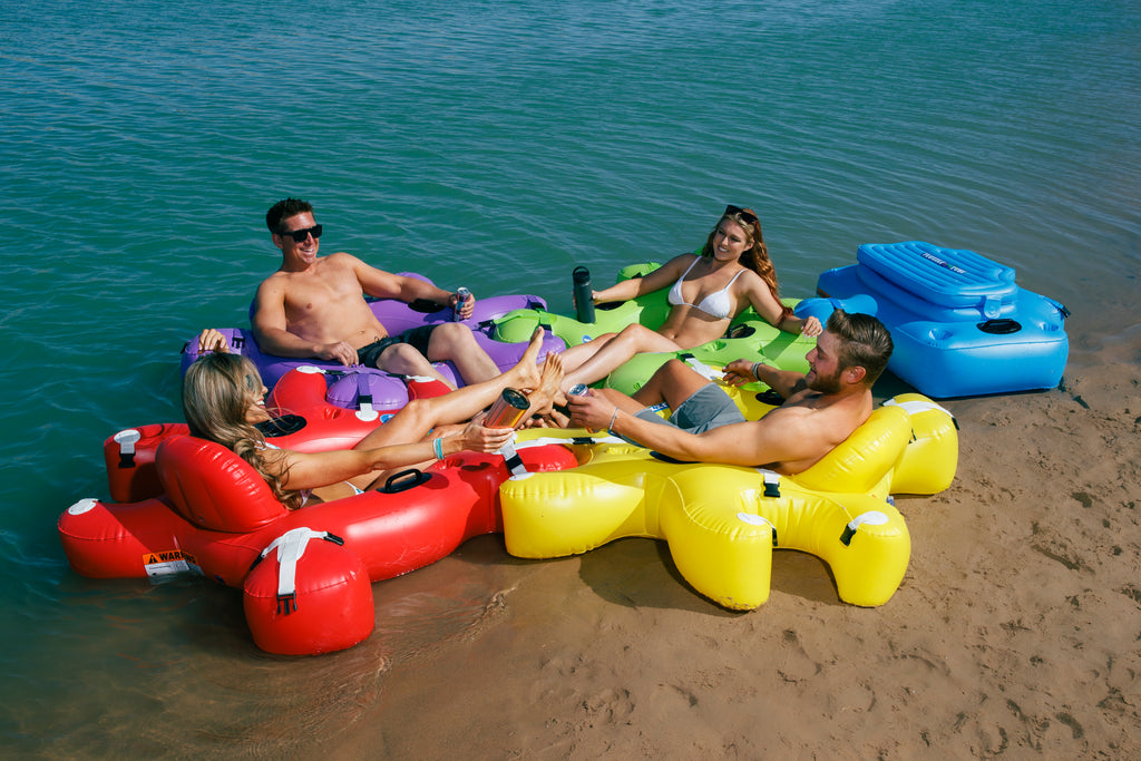 4 people in bathing suite floating in Red, Green, Purple and Yellow Fluzzle Tubes puzzle shaped inflatable and interlocking river, lake or pool tube with inflatable back rest, mesh bottom, expandable cup holders, 2 durable handles. 12 nylon connectors. Interlocking floating cooler. 