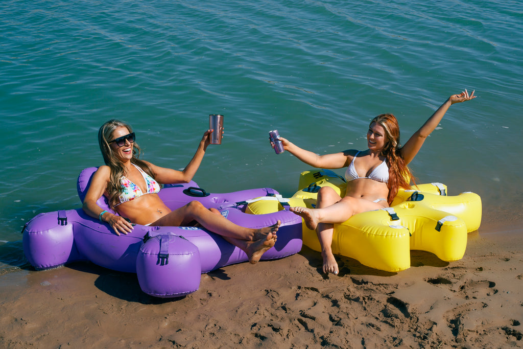 Two woman floating in Purple and Yellow Fluzzle Tubes- puzzle shaped inflatable and interlocking river, lake or pool tube with inflatable back rest, mesh bottom, expandable cup holders, 2 durable handles. . 12 nylon connectors