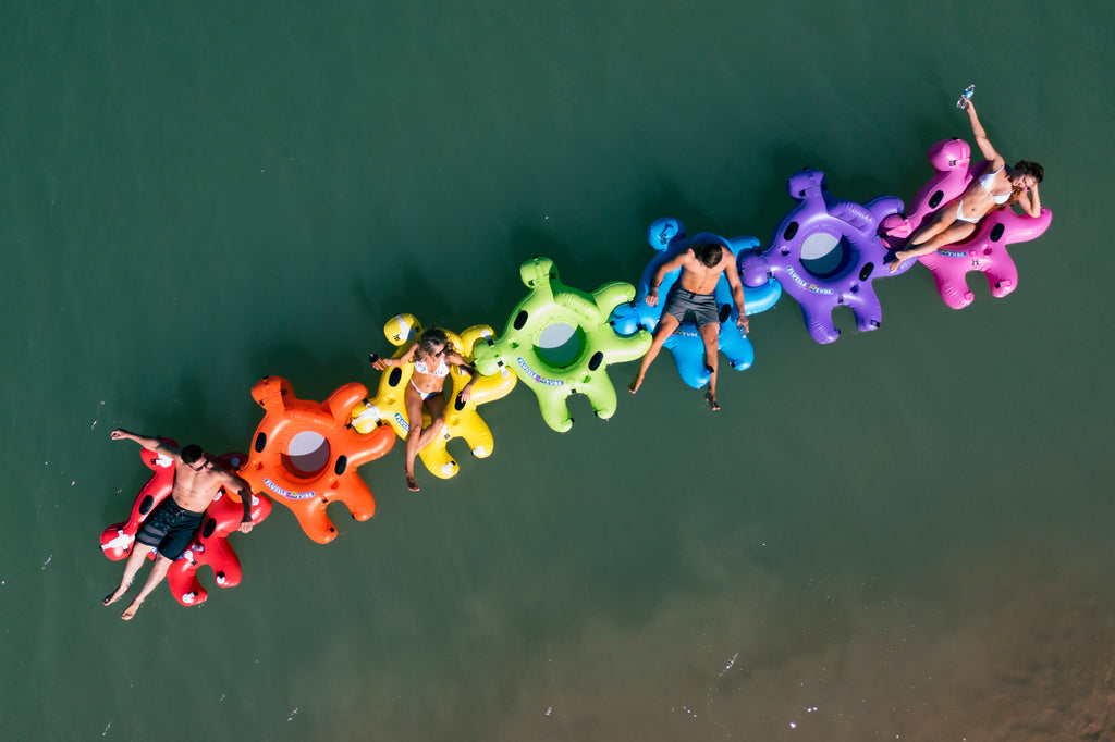 4 people in bathing suite floating in Red, Green, Purple and Yellow Fluzzle Tubes puzzle shaped inflatable and interlocking river, lake or pool tube with inflatable back rest, mesh bottom, expandable cup holders, 2 durable handles. 12 nylon connectors. Interlocking floating cooler.  Rainbow collection.  Rainbow inflatable tubes for river, lake or pool.